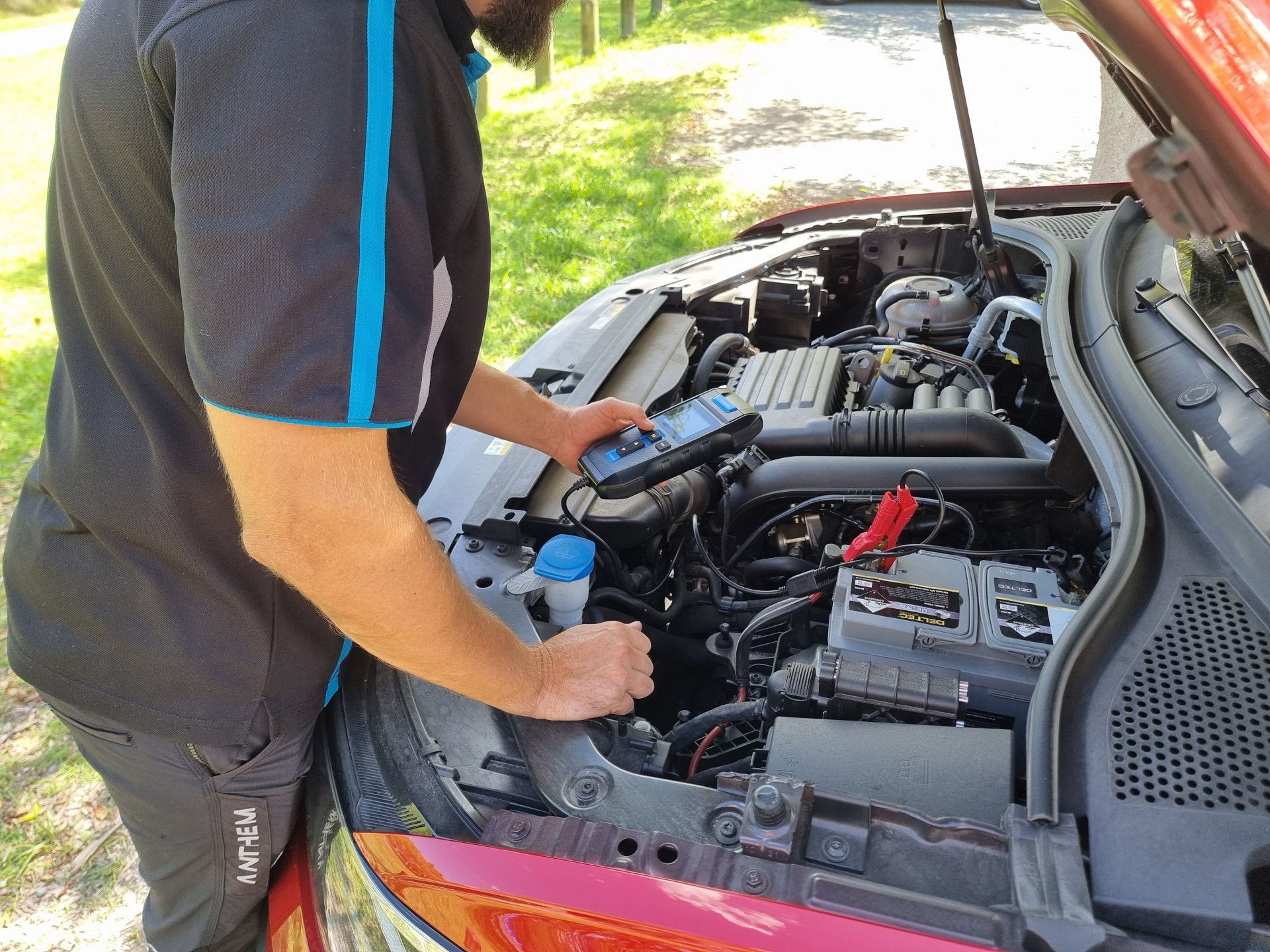 Technician testing a car battery with a diagnostic tool under the bonnet of a red vehicle
