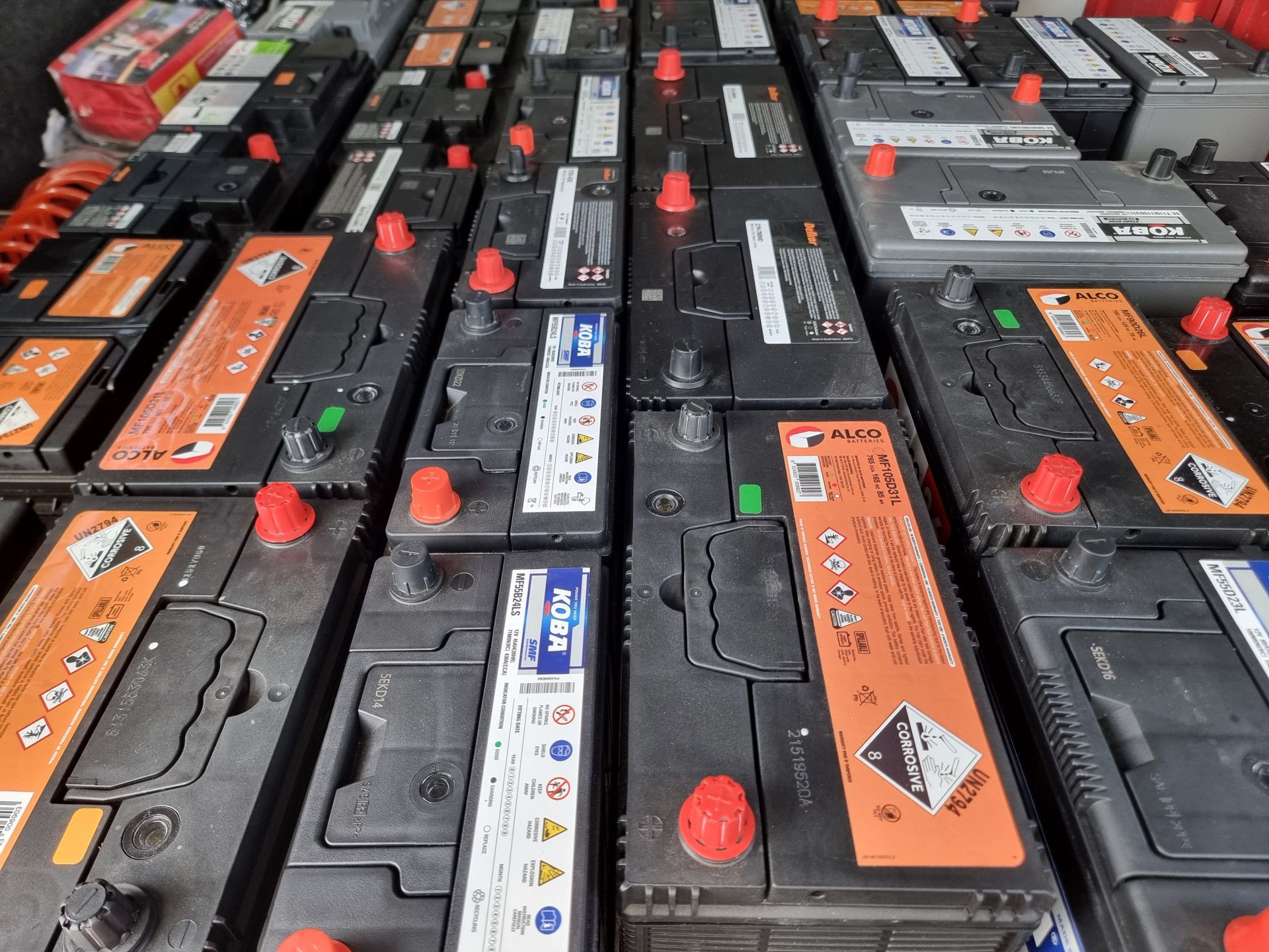 Rows of car batteries in storage, featuring multiple brands with red terminal caps and hazard labels