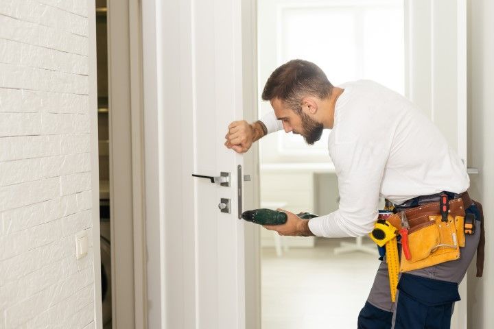 Man in a white shirt and tool belt installing a door handle with a power drill in a white interior.