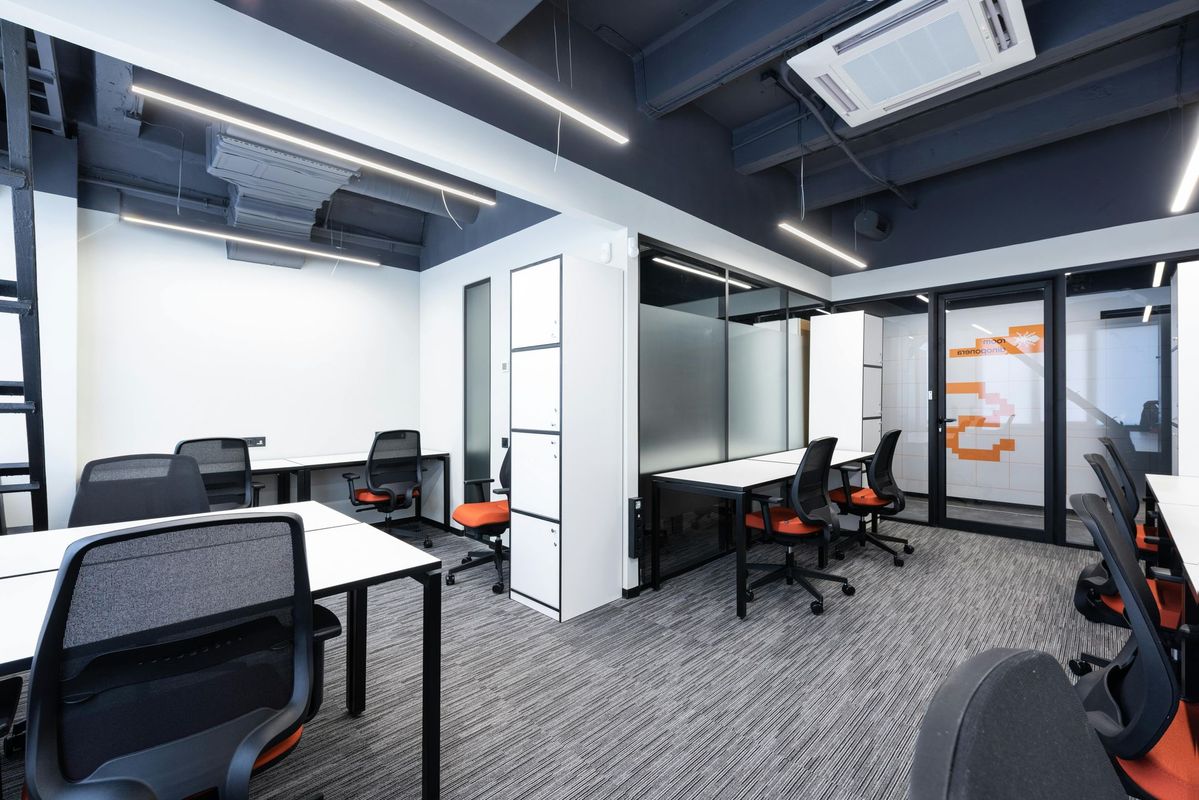 Modern open office with desks, black chairs, glass partitions, and gray carpet under bright ceiling lights