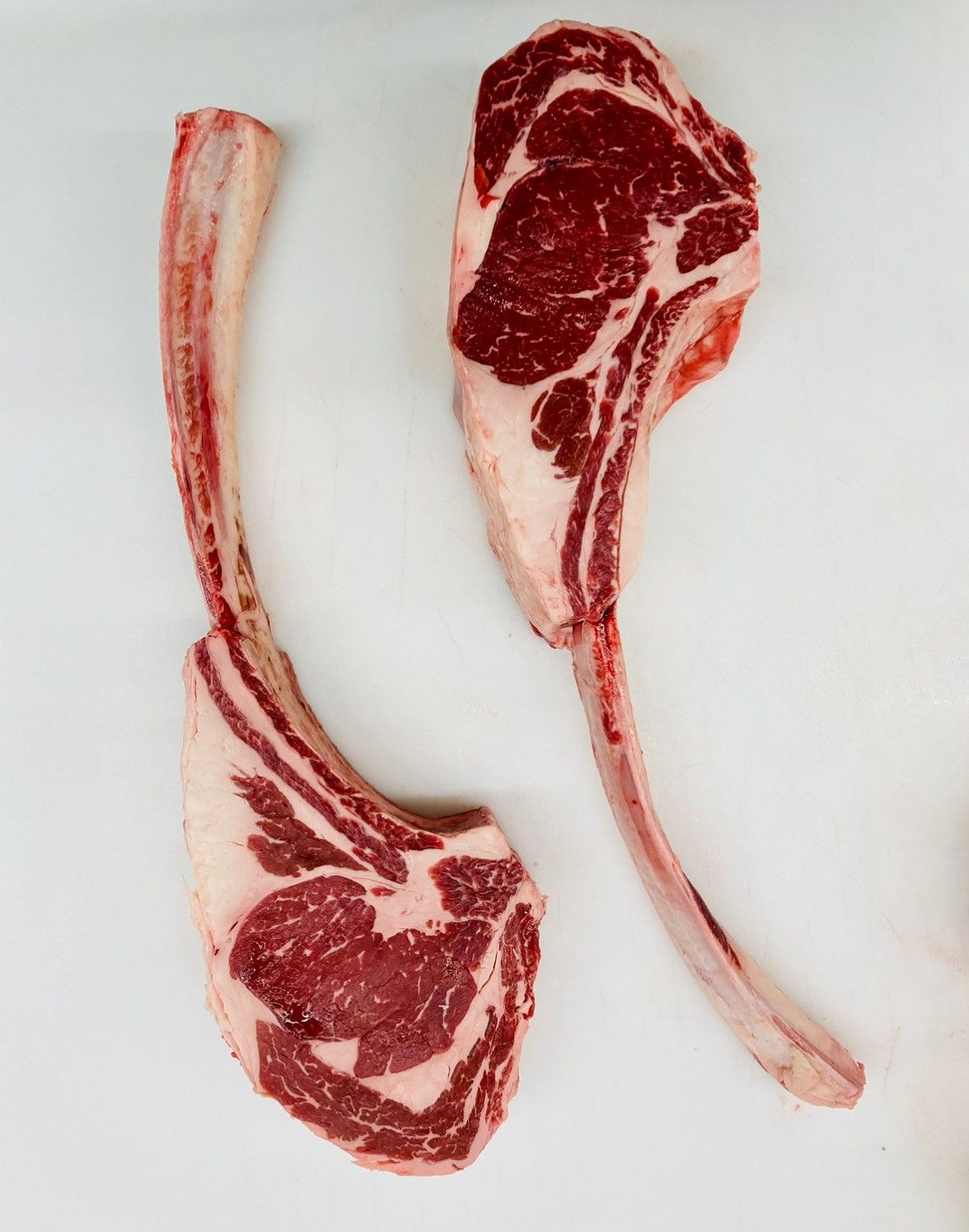 Two raw, marbled tomahawk steaks with long, exposed rib bones placed against a plain white background.