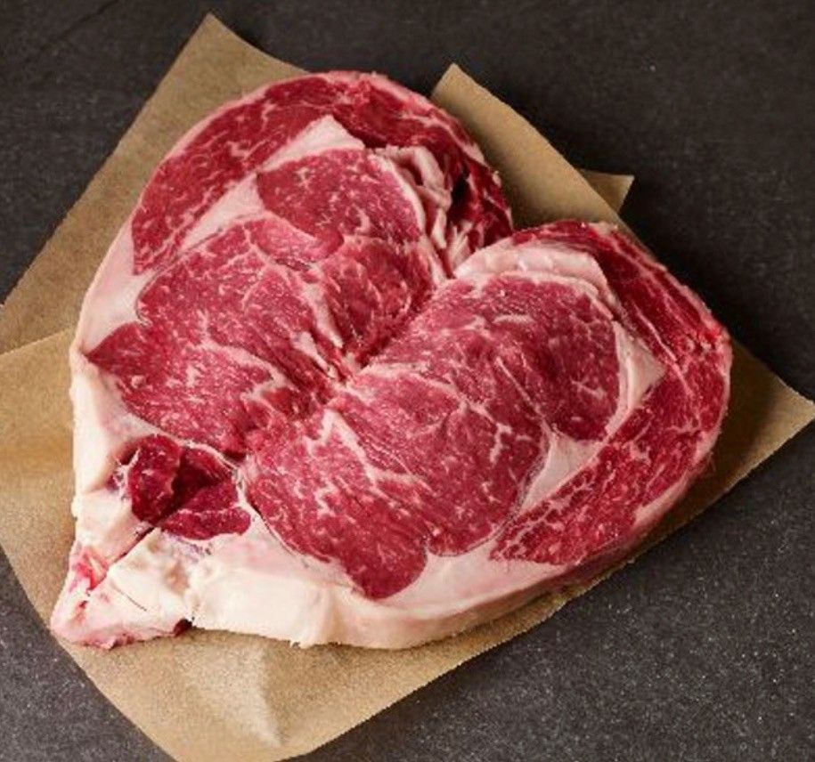 A raw, marbled ribeye steak sitting on a piece of brown butcher paper against a dark, textured surface.