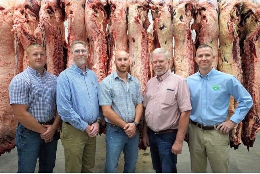 Five people stand in a line posing for a photo in a refrigerated room with hanging beef carcasses behind them.