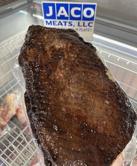 A large, vacuum-sealed smoked brisket displayed in a refrigerated case with a Jaco Meats LLC sign in the background.