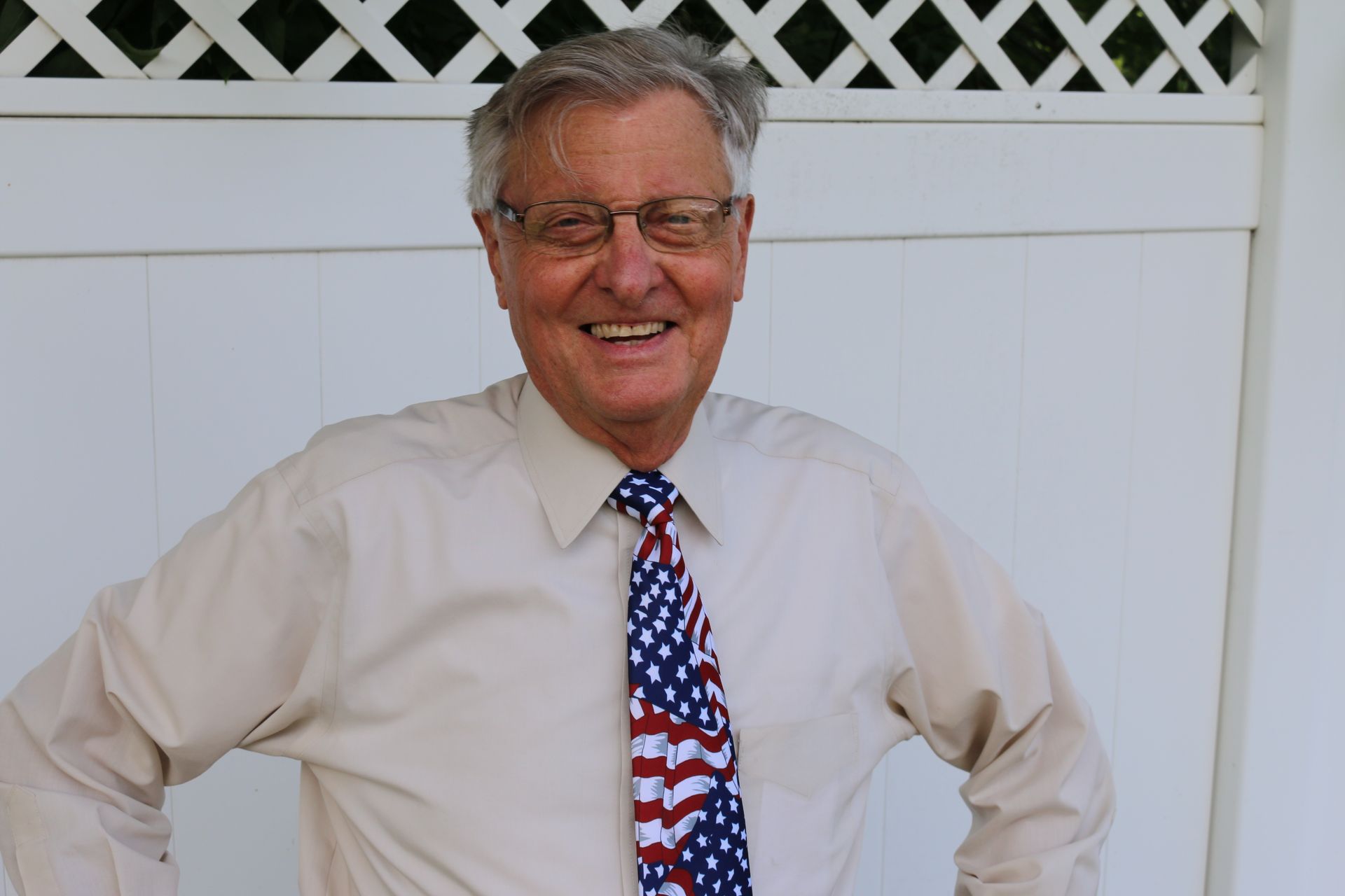 Smiling man in glasses wearing a white shirt and American flag tie, standing in front of a white fence.