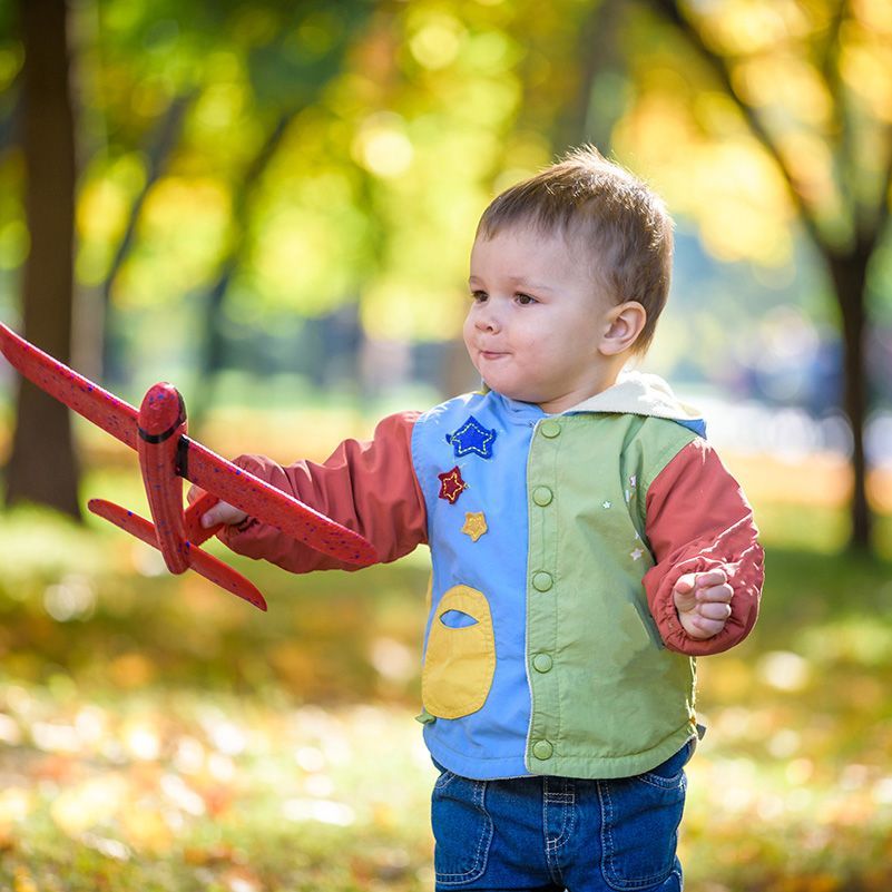Toddler in a green vest holds a red toy airplane in a sunny park