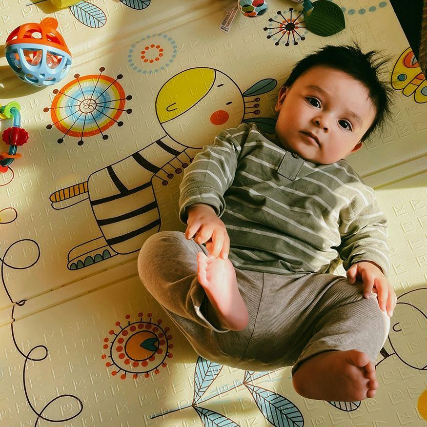 Baby lying on a patterned play mat, reaching toward a colorful toy ball.