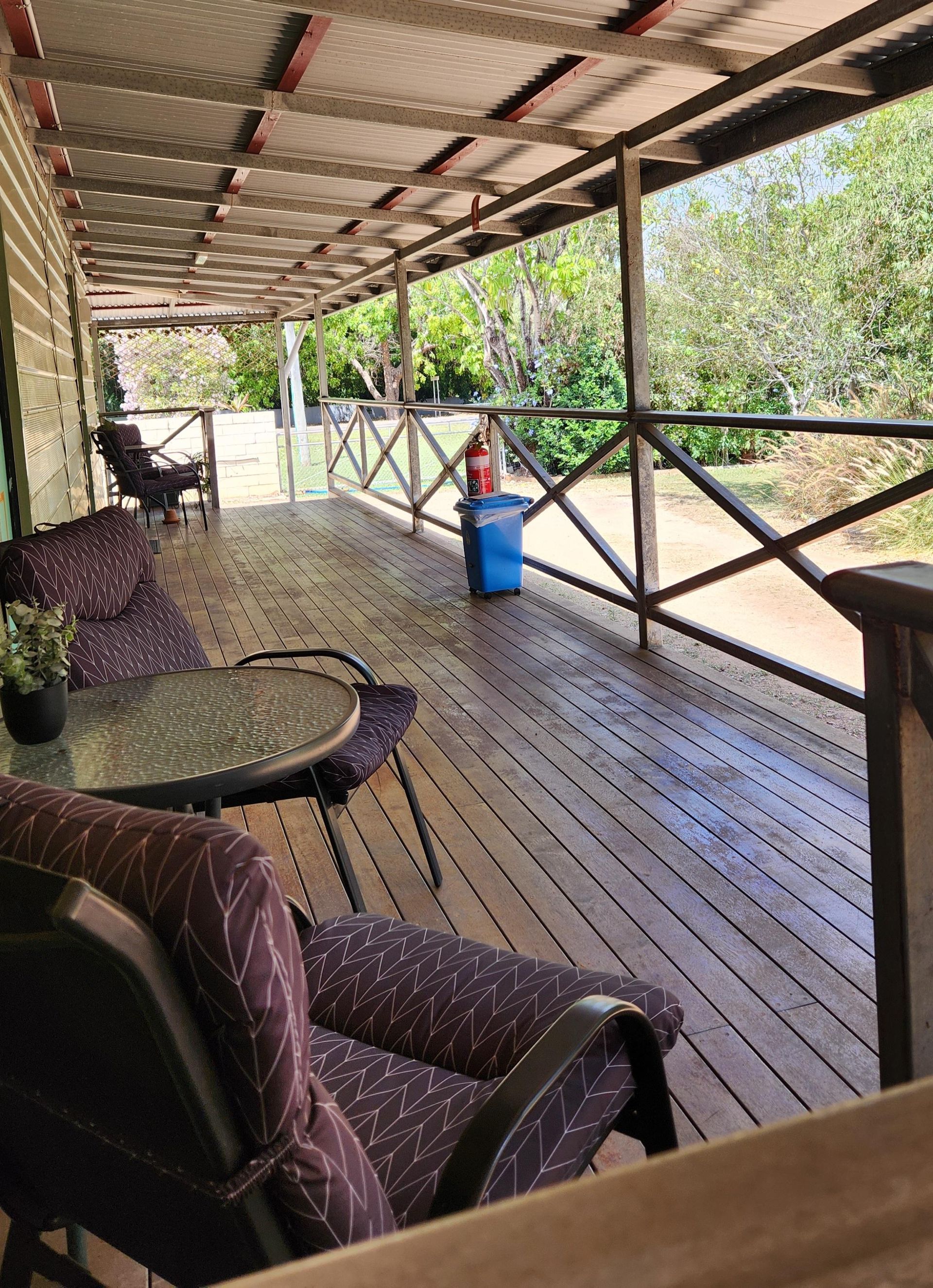 Covered wooden porch with chairs, table, blue barrel, and railing overlooking trees and a dirt driveway
