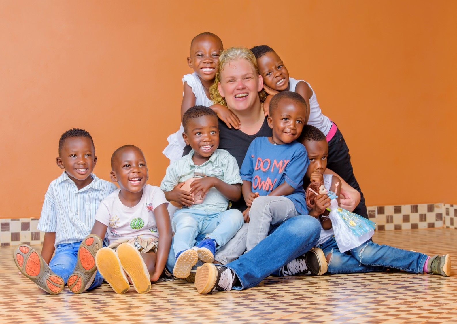 Een vrouw glimlacht, omringd door lachende kinderen, op een tegelvloer tegen een oranje muur.