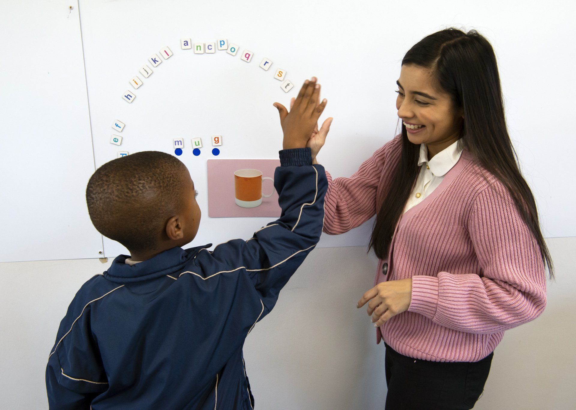 A young boy high-fives a smiling woman near a whiteboard in a classroom.