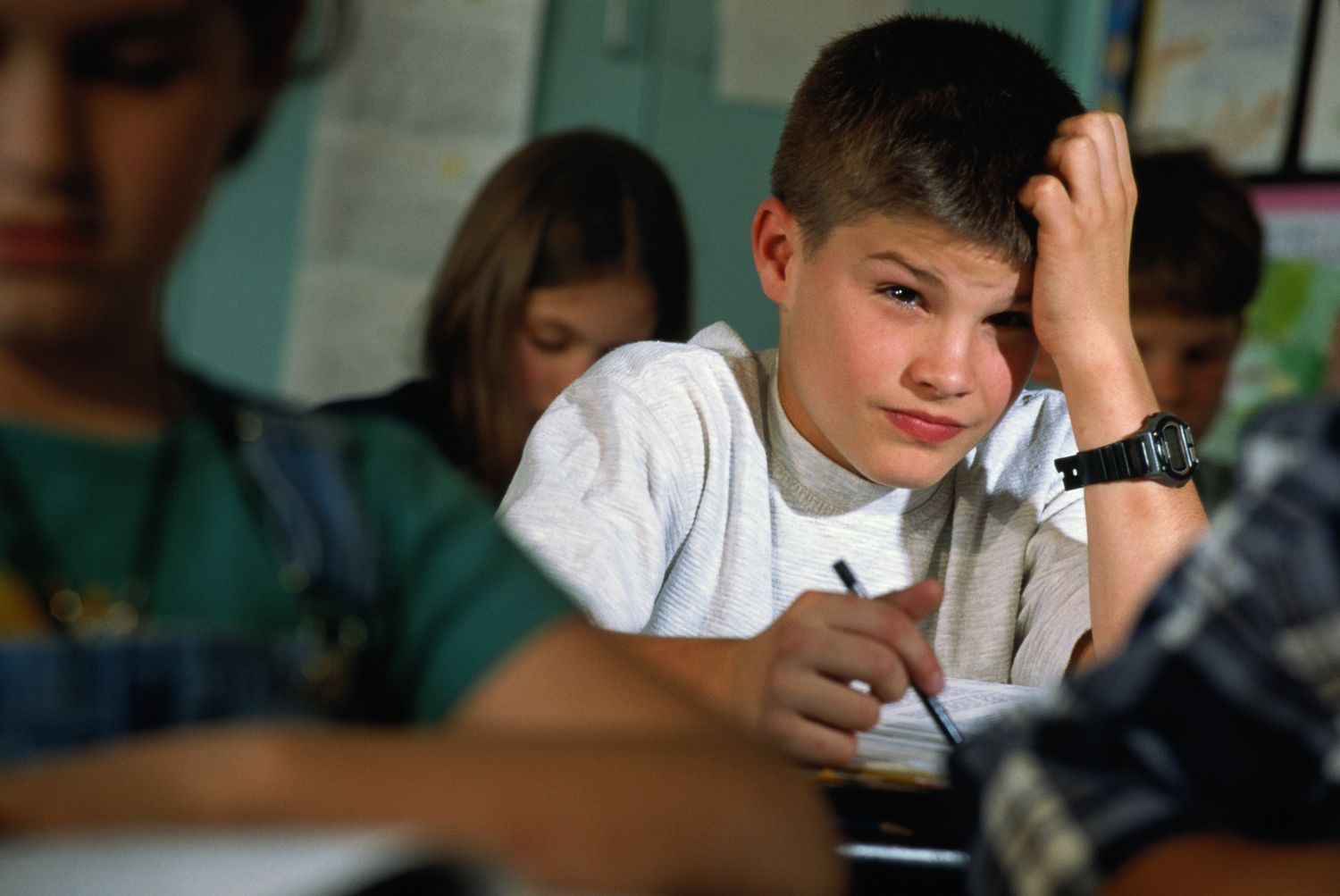 A boy is sitting at a desk in a classroom with his hand on his head.