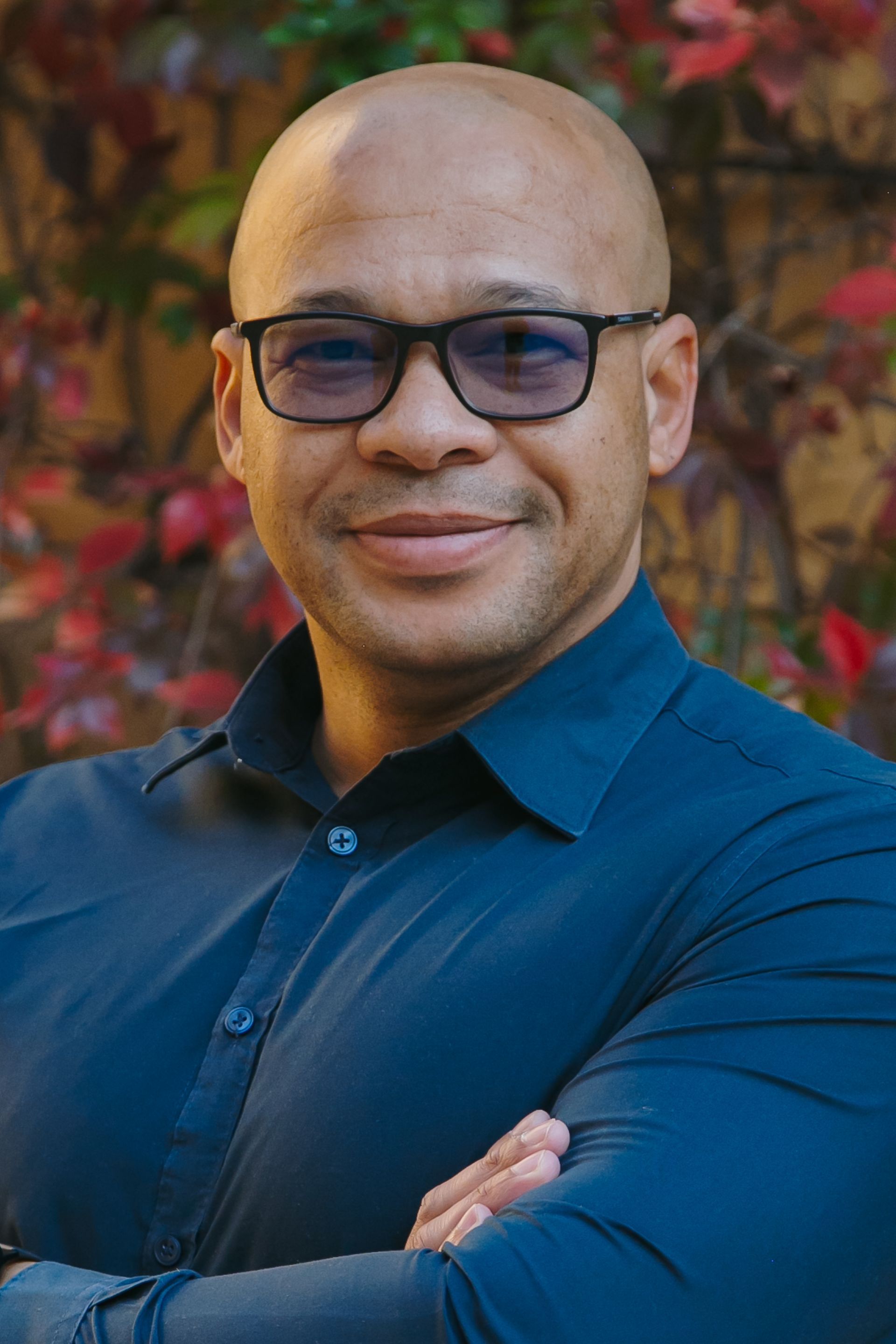 A bald man wearing glasses and a blue shirt is standing with his arms crossed.
