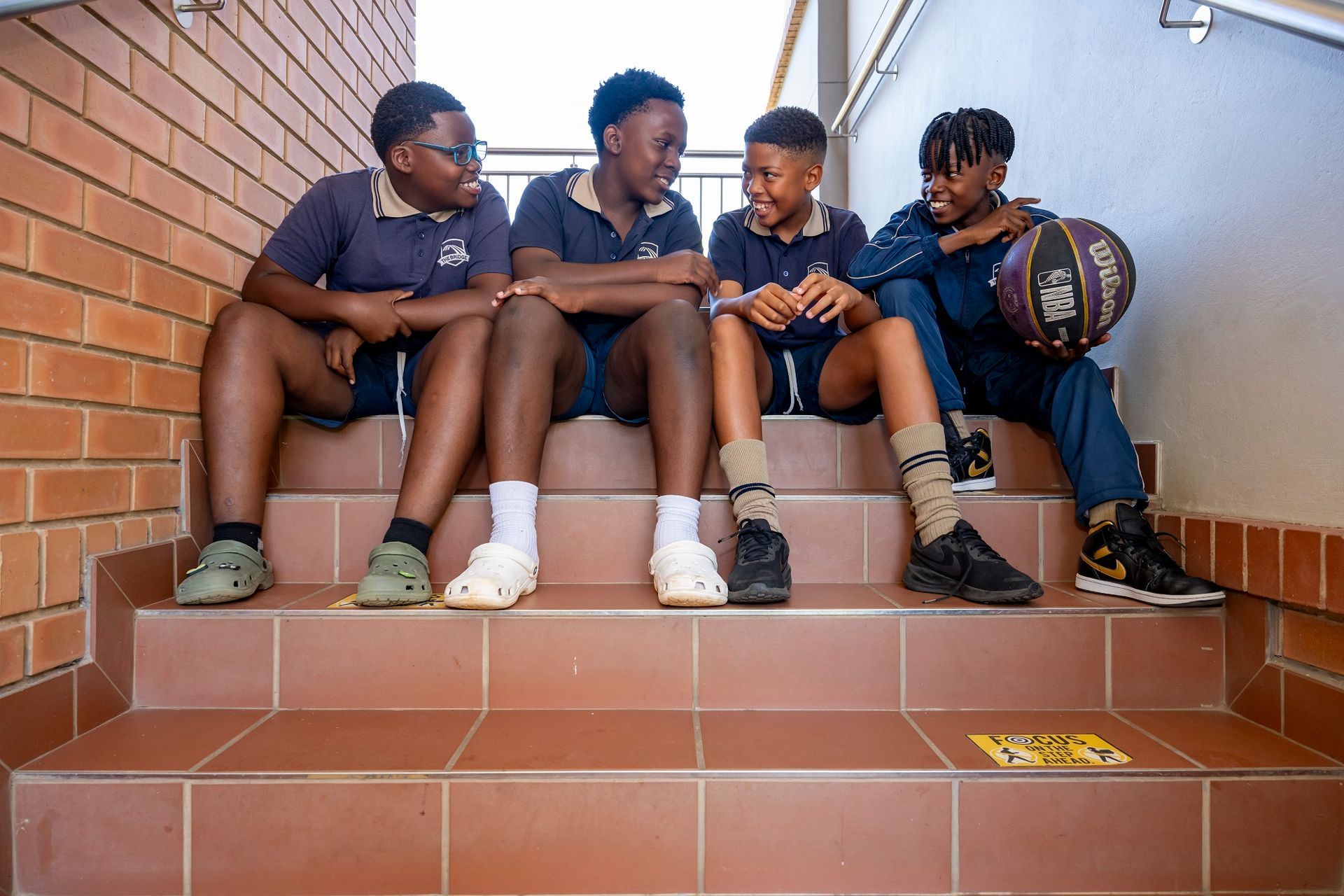Four male students in school uniforms sit on stairs, talking, one holding a basketball.