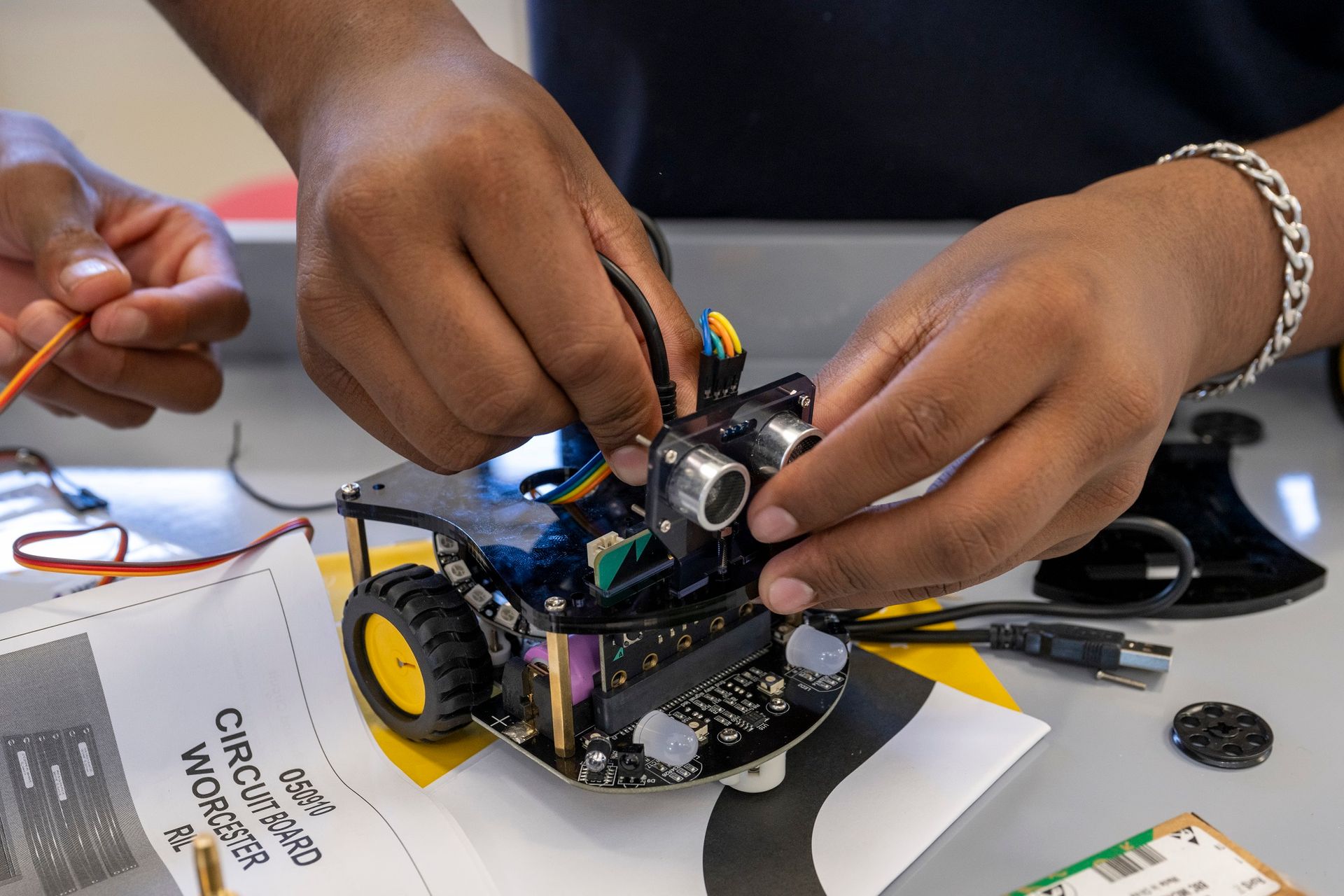 Hands assembling a small robot on a table with instructions and electronic components.