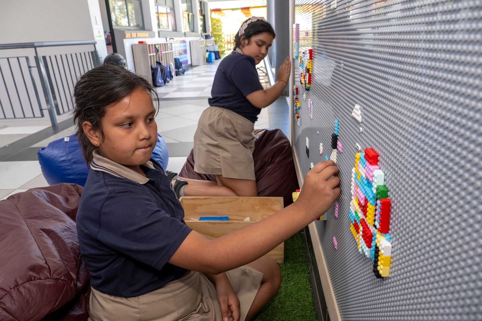 Two girls building with Legos on a wall at school. One is seated, the other standing.