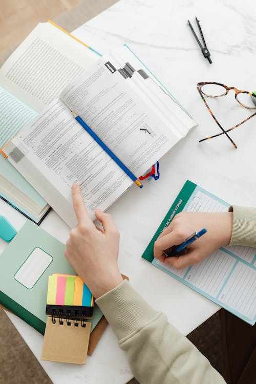 A person is sitting at a desk reading a book and writing in a notebook.