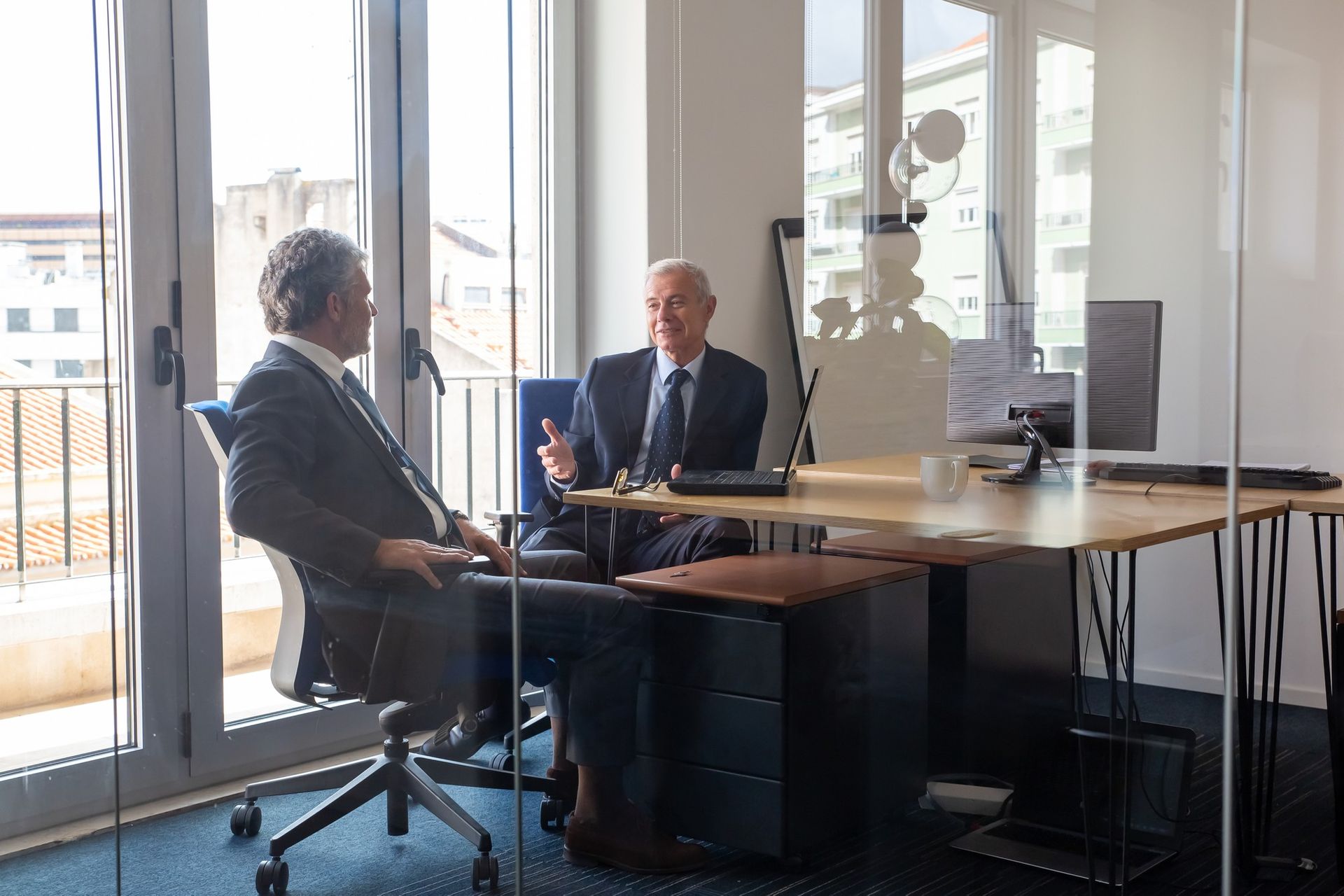 Two people in suits meeting across a table in a glass-walled office