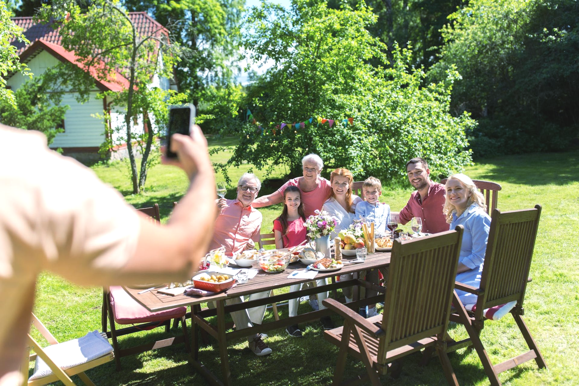 Friends gathered around an outdoor dining table in a sunny garden while someone takes a photo.