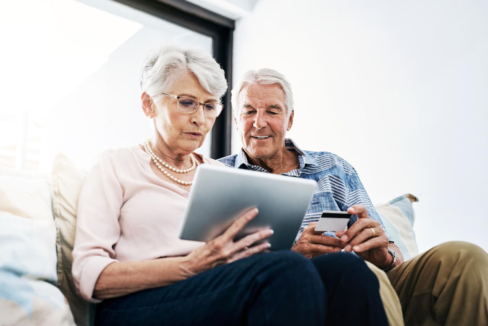 Older couple sitting on a couch, looking at a tablet and credit card in a bright living room.