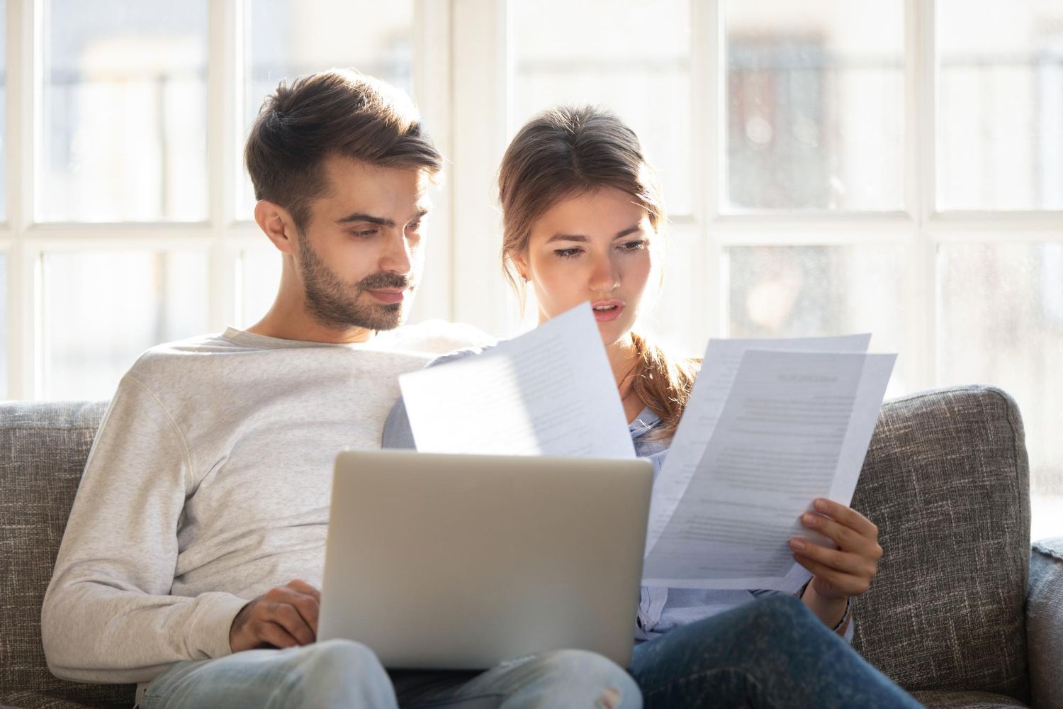Two people reviewing papers and working on a laptop on a couch in a bright living room
