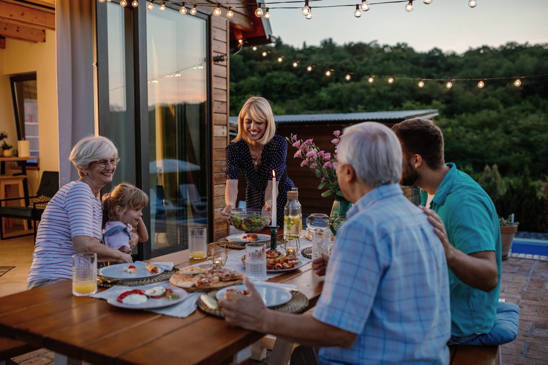 Friends sharing dinner on a lit patio at dusk, with plates, drinks, and string lights overhead.