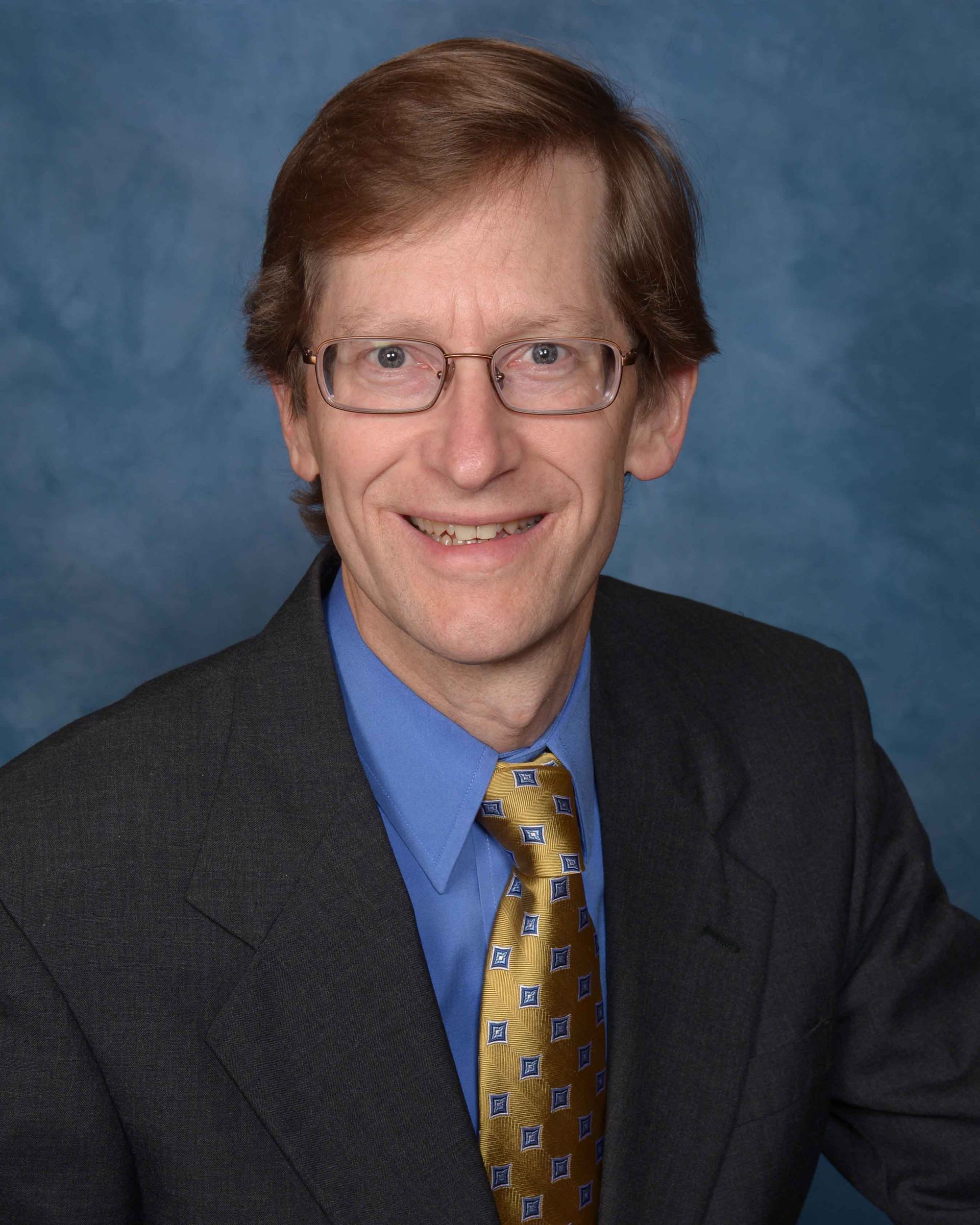 Portrait of a smiling man in a dark suit, blue shirt, and patterned gold tie against a blue backdrop