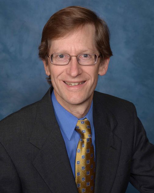 Portrait of a smiling man in a dark suit, blue shirt, and patterned gold tie against a blue backdrop