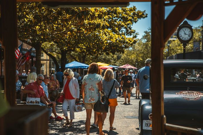 Crowded sunny street market with shoppers, trees, umbrellas, and hanging signs