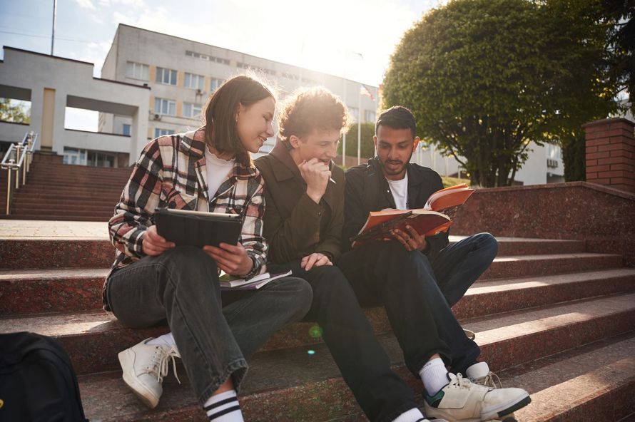 Three students sit on campus steps, talking and reading notebooks in the sunlight.