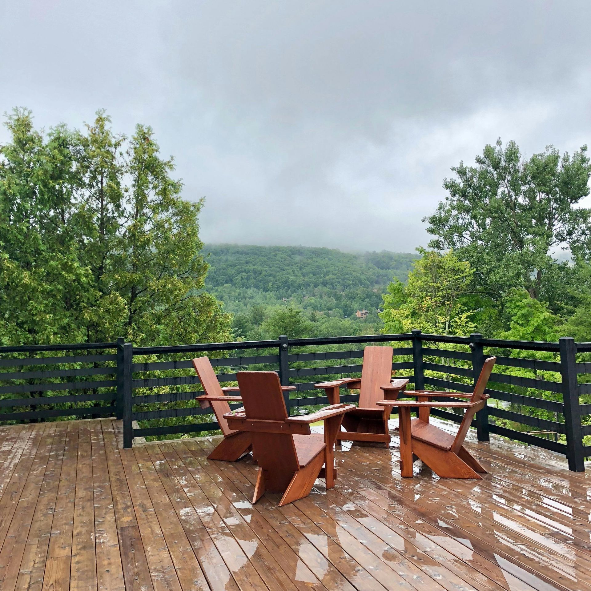 Wet wooden deck with Adirondack chairs overlooking a misty green mountainside.