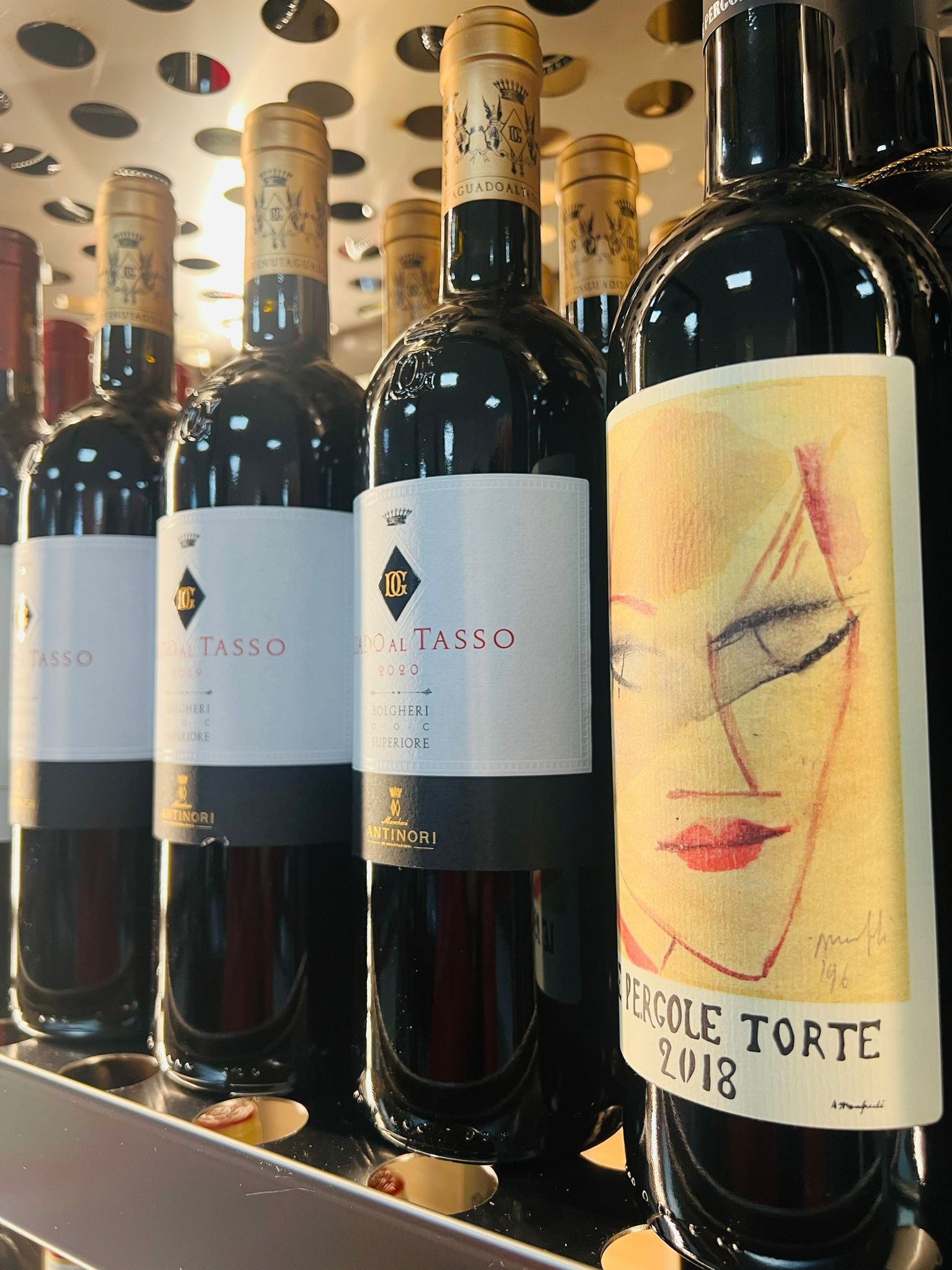 Su uno scaffale si trovano bottiglie di vino rosso, tra cui una Le Pergole Torte 2018 con un'etichetta caratterizzata da un'originale illustrazione astratta di un volto.
