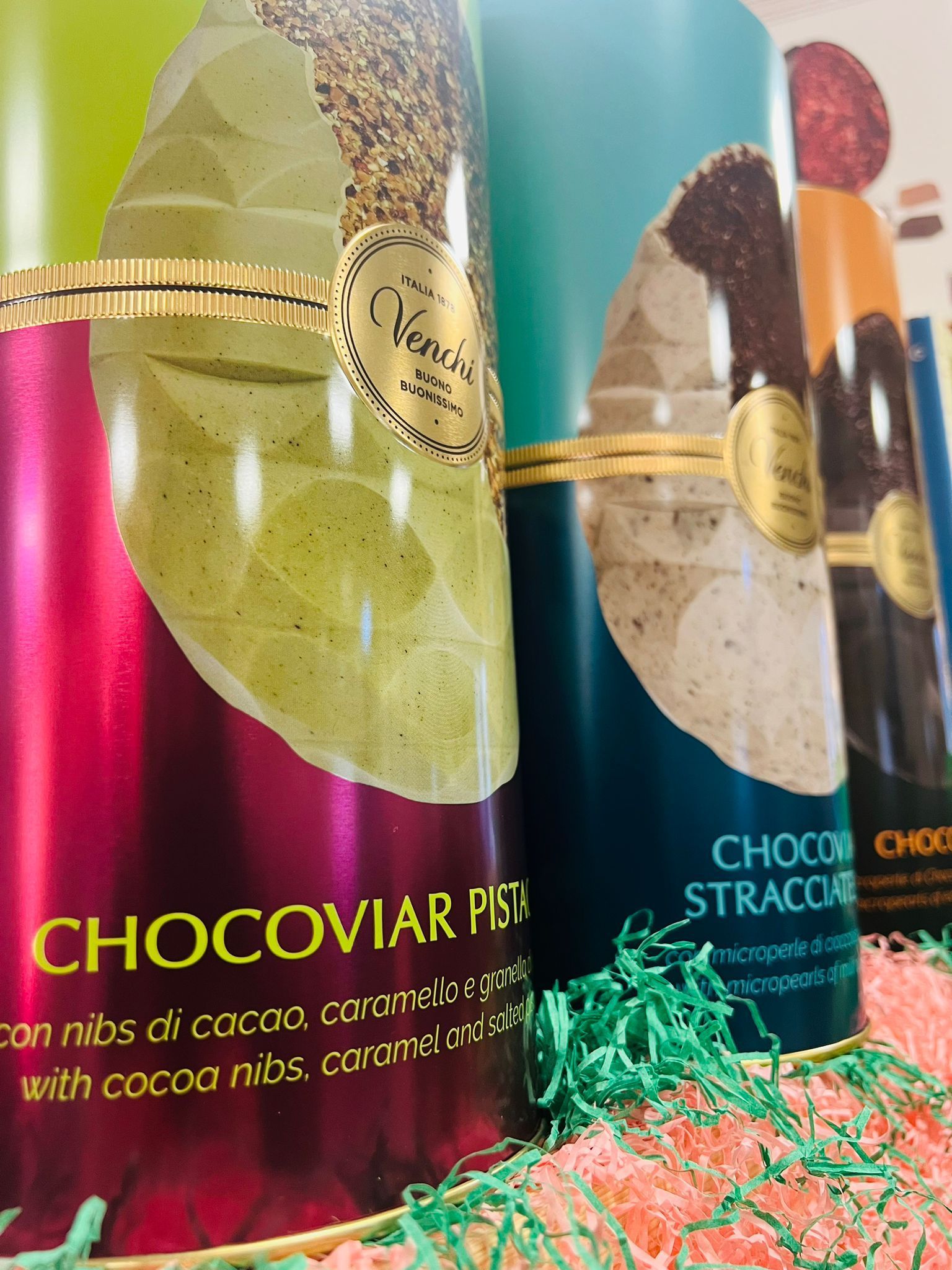 Tre scatole cilindriche di cioccolatini Venchi, nei colori verde, turchese e arancione, poggiate su un letto di erba decorativa verde e rosa.