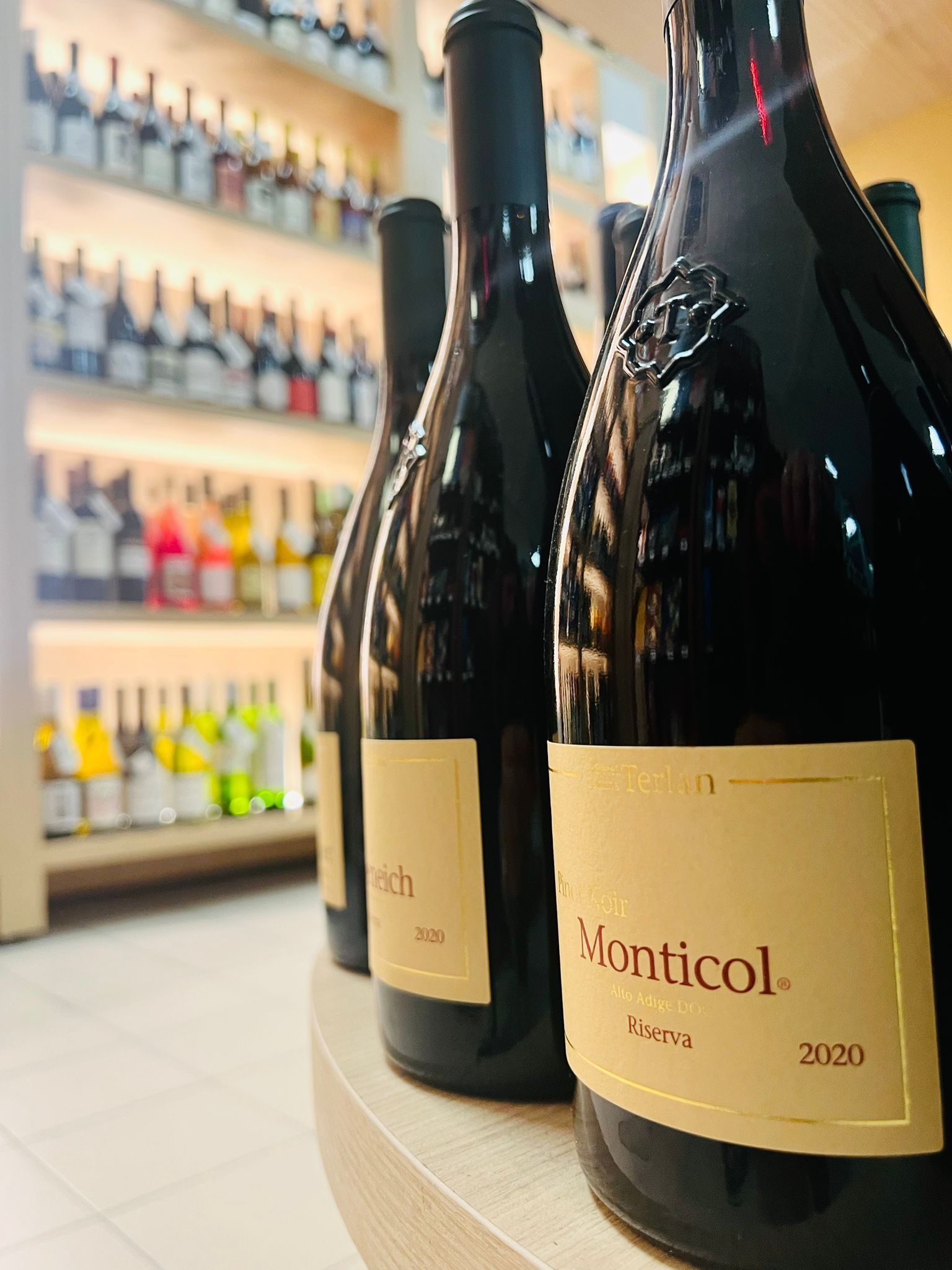 In primo piano, bottiglie di vino rosso Monticol, mentre sullo sfondo si intravedono scaffali con diverse bottiglie di vino, sfocati.