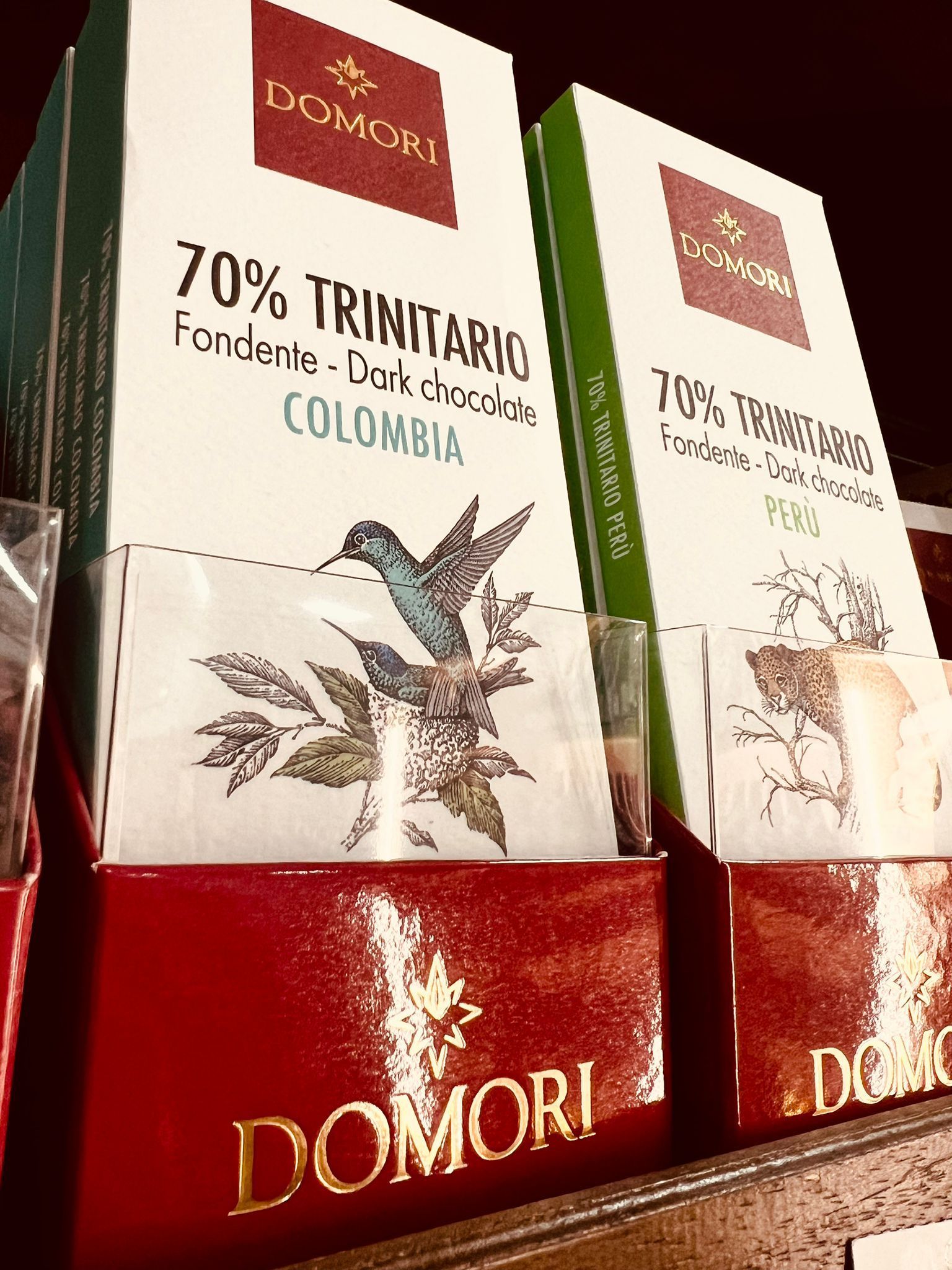 Due tavolette di cioccolato Domori con confezione bianca, decorate con l'illustrazione di un colibrì e del testo, esposte su uno scaffale rosso.
