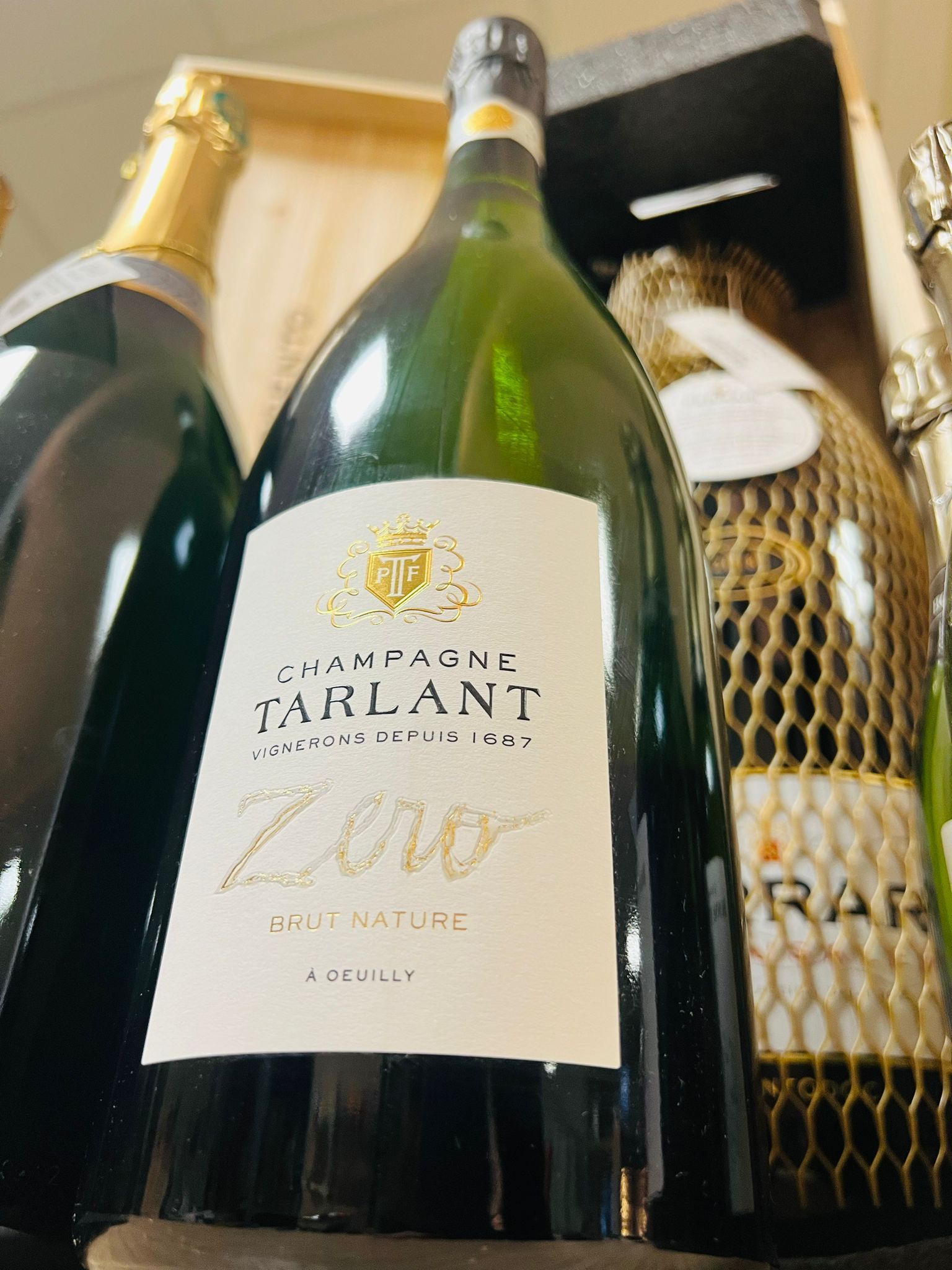 Una bottiglia di champagne Jarlant Zero è posizionata al centro di un espositore, con altre bottiglie di vino parzialmente visibili ai lati.