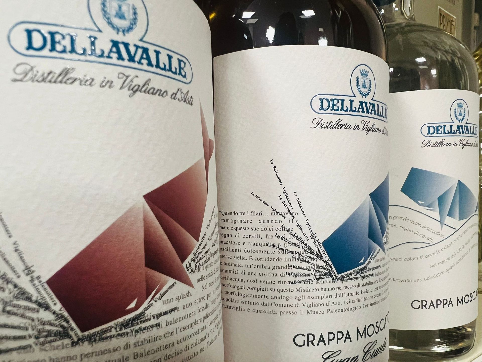 Su uno scaffale sono allineate tre bottiglie di grappa del marchio Dellavalle, ognuna con una particolare grafica in stile origami.