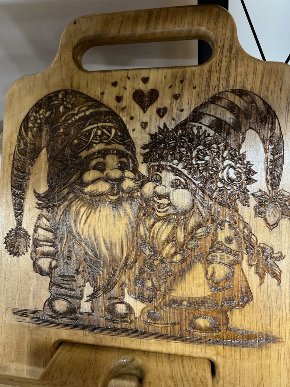 Wooden cutting board with carved gnome and witch under hearts and leaves, stained in dark brown.