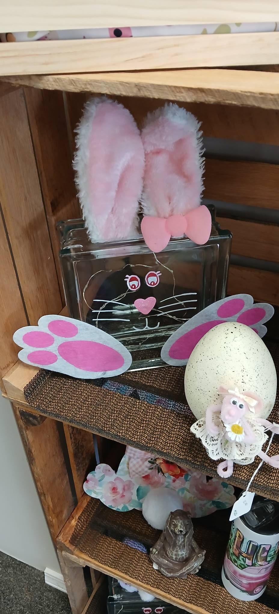 Pink bunny ears and paw print decorations on a small shelf display, with a white egg ornament and jars below.