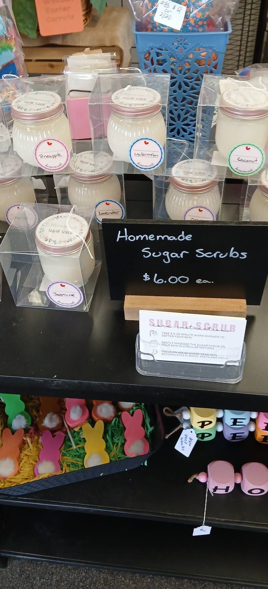 Display of homemade sugar scrubs in small jars with a black price sign on a shelf.