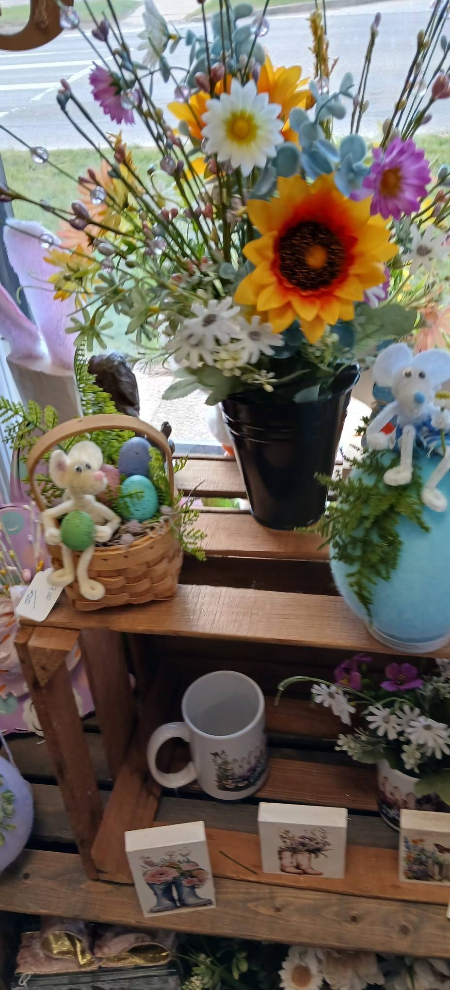 Flower bouquet in a vase on a wooden shelf, surrounded by small figurines, mugs, and framed photos.