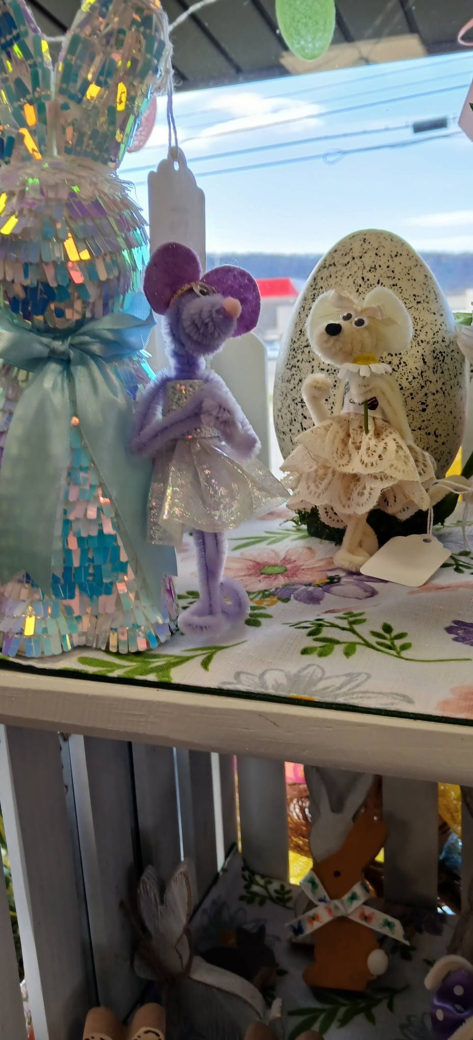 Small pastel figurines and a crocheted doll displayed in a shop window with decorative lights and shelves