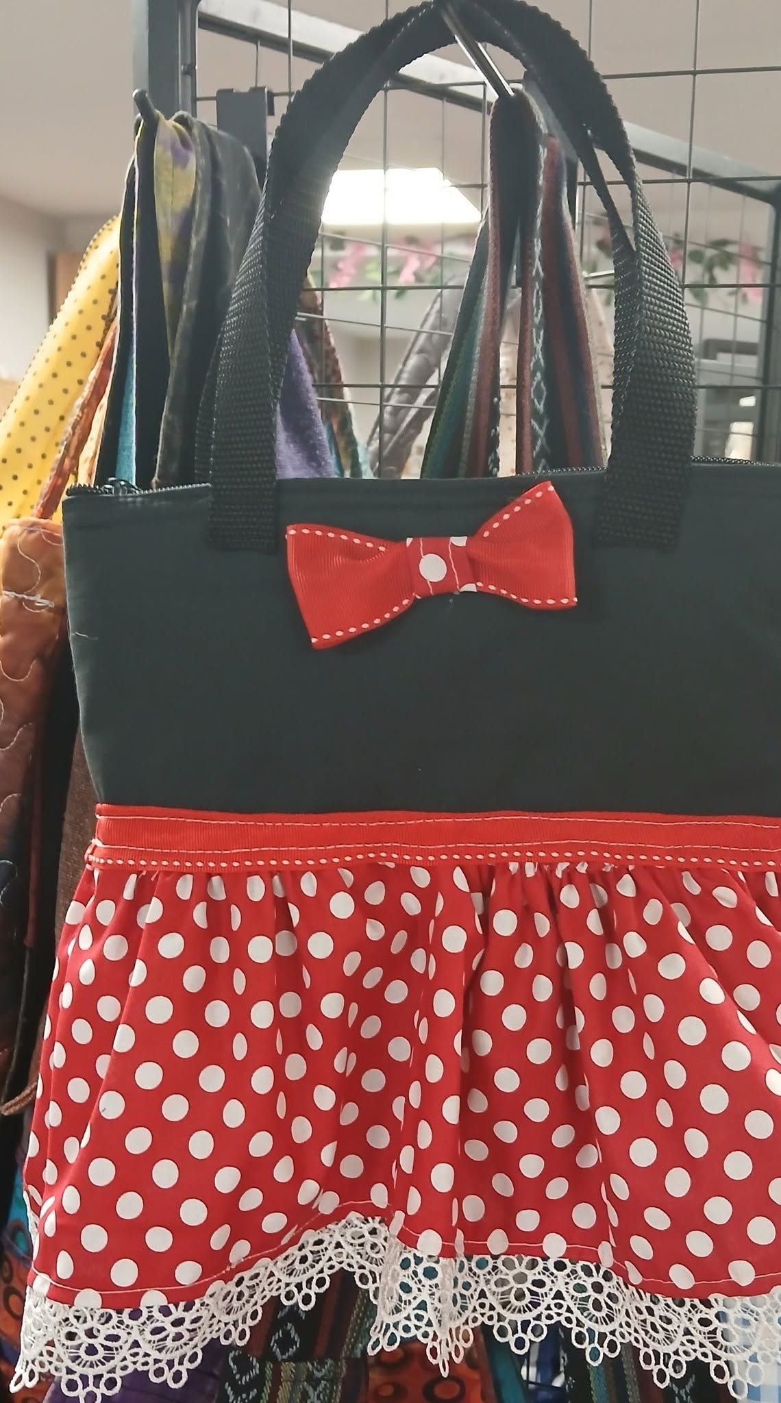 Black tote bag with a red bow and red polka-dot ruffle hanging on a hook, indoors.