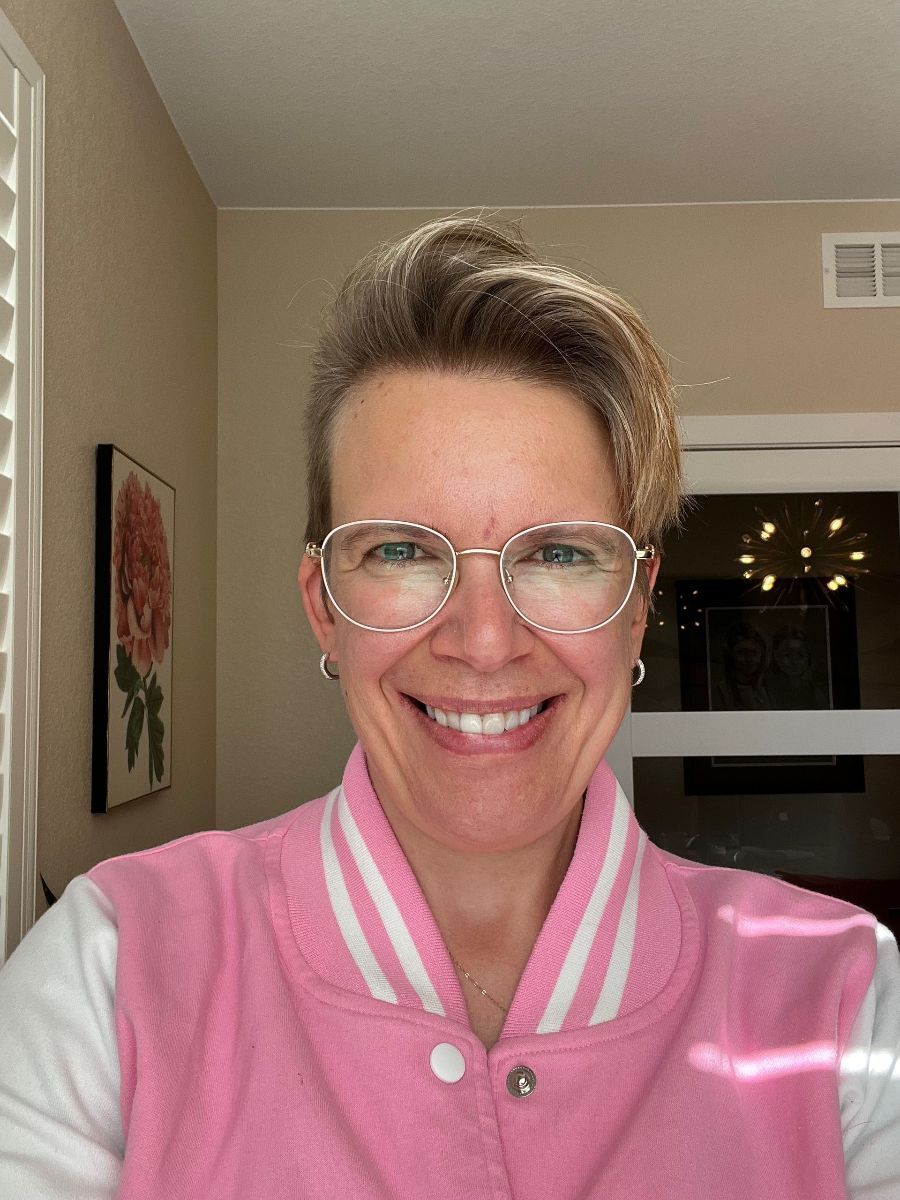 Smiling person in a pink varsity jacket and clear glasses indoors, with a framed flower print in the background
