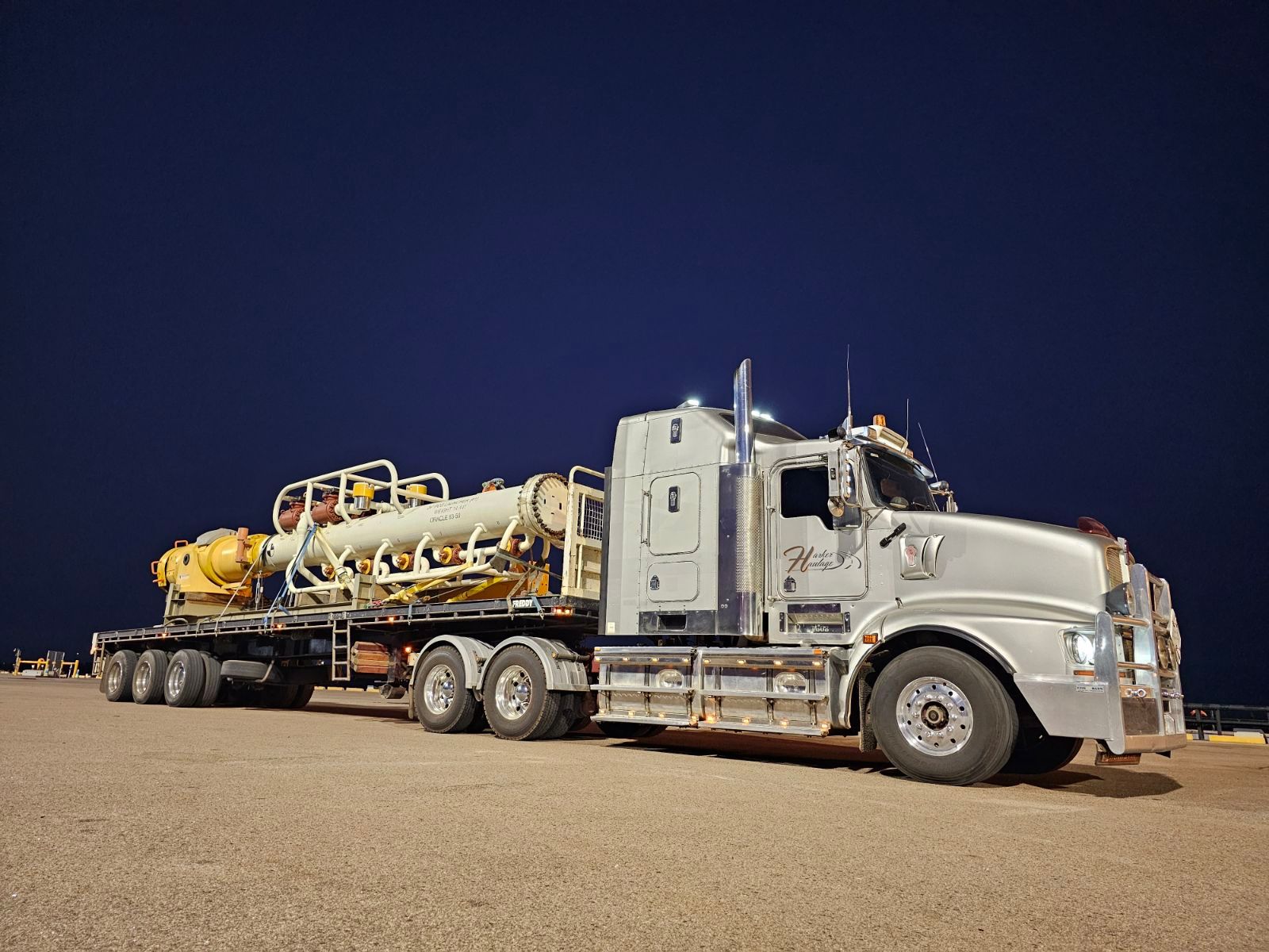Silver semi-truck with oversized equipment on a flatbed trailer on a gravel lot at night. — Harker Haulage In East Arm, NT