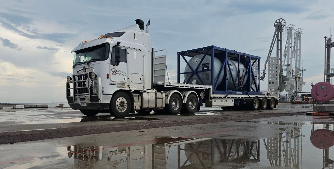 A white truck with a large blue cage carrying a metallic tank on a dock near crane infrastructure. — Harker Haulage In East Arm, NT