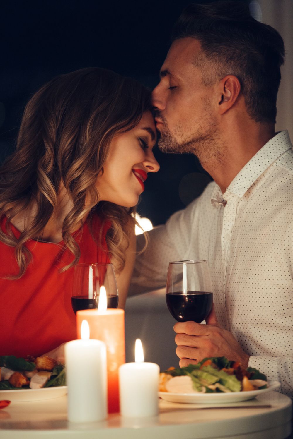 Un hombre besa a una mujer en la frente durante una cena a la luz de las velas. Sostienen copas de vino y un vestido rojo en un ambiente romántico.