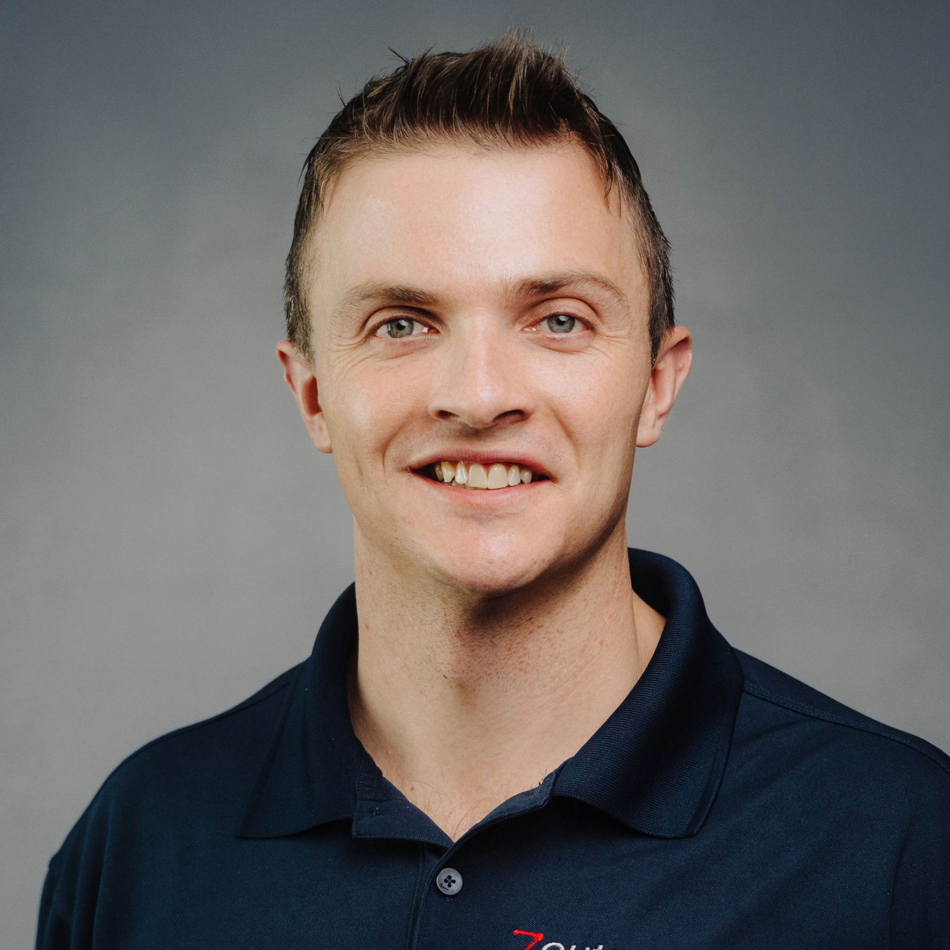 Smiling man with short brown hair, wearing a blue polo shirt. Gray background.