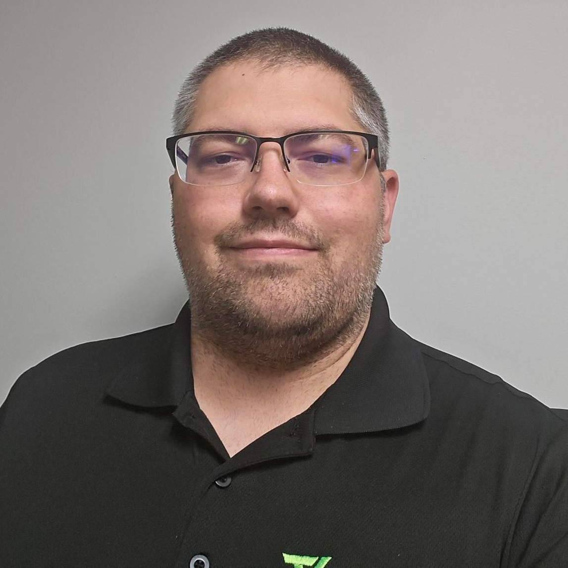 Man with glasses, smiling, wearing a black polo shirt, in front of a grey wall.