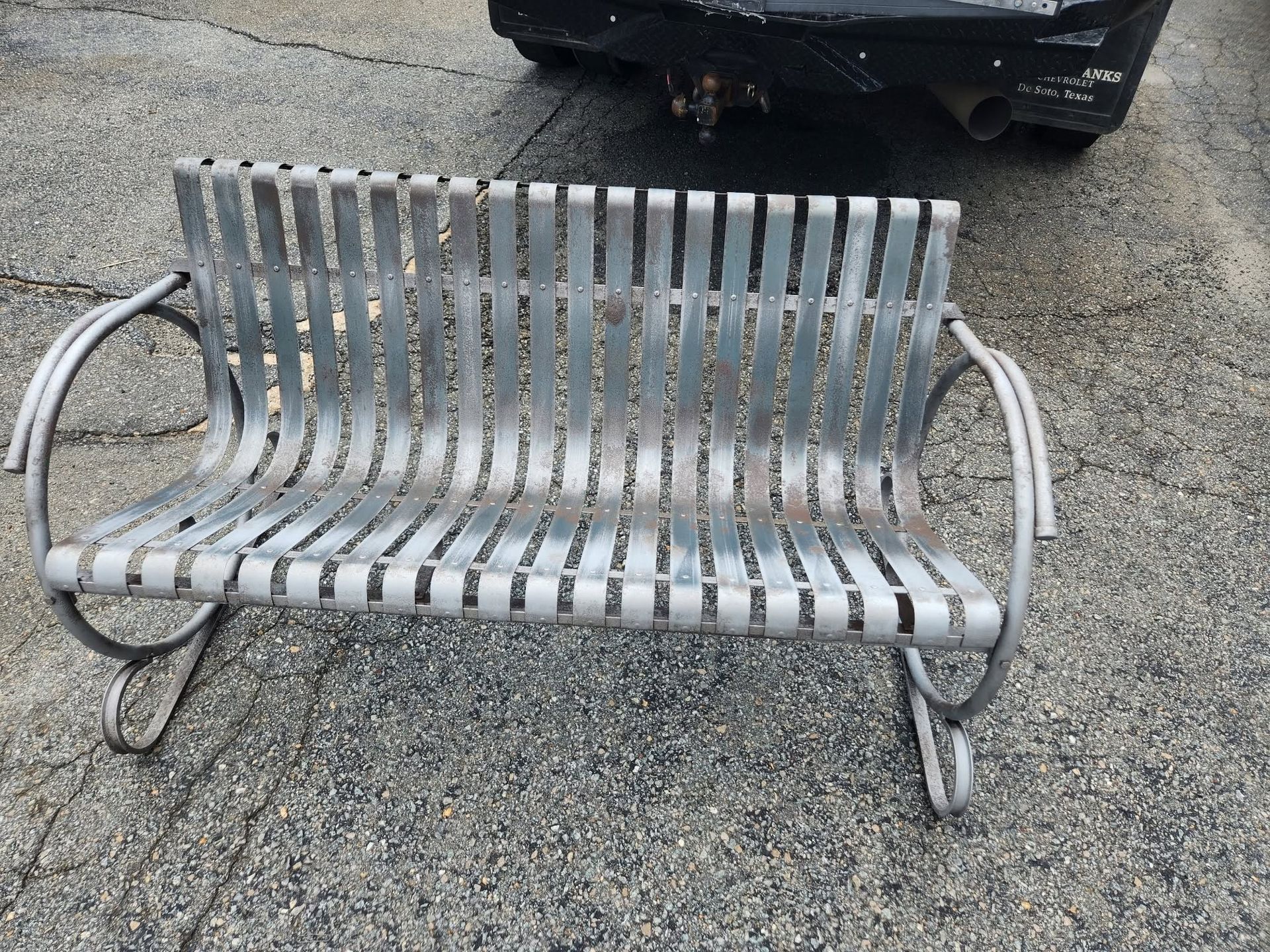 Weathered metal bench with slatted seat and backrest on a gravel parking lot, with a car behind it