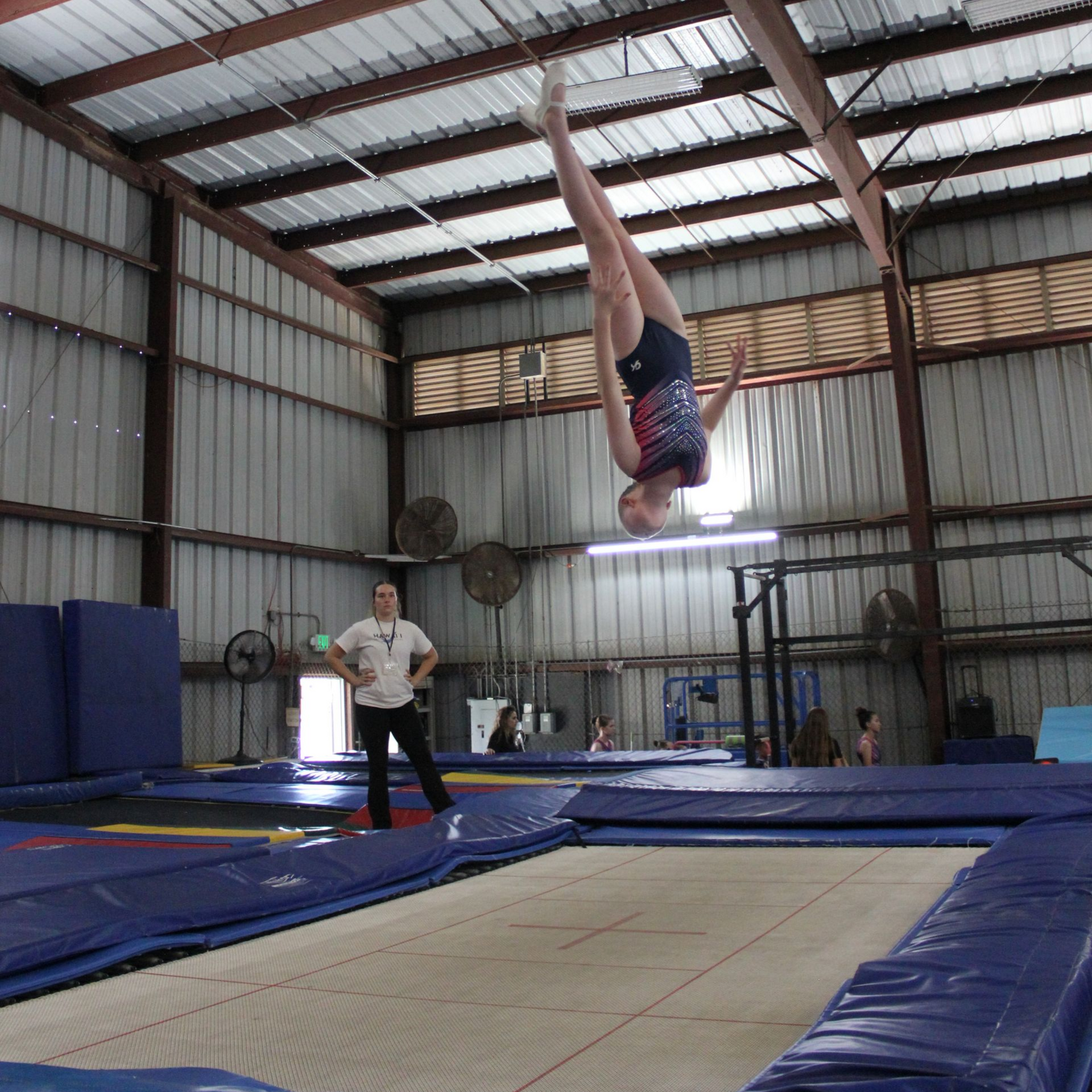 Girl in blue and purple clothing upside down on a trampoline with instructor spotting with a mat.